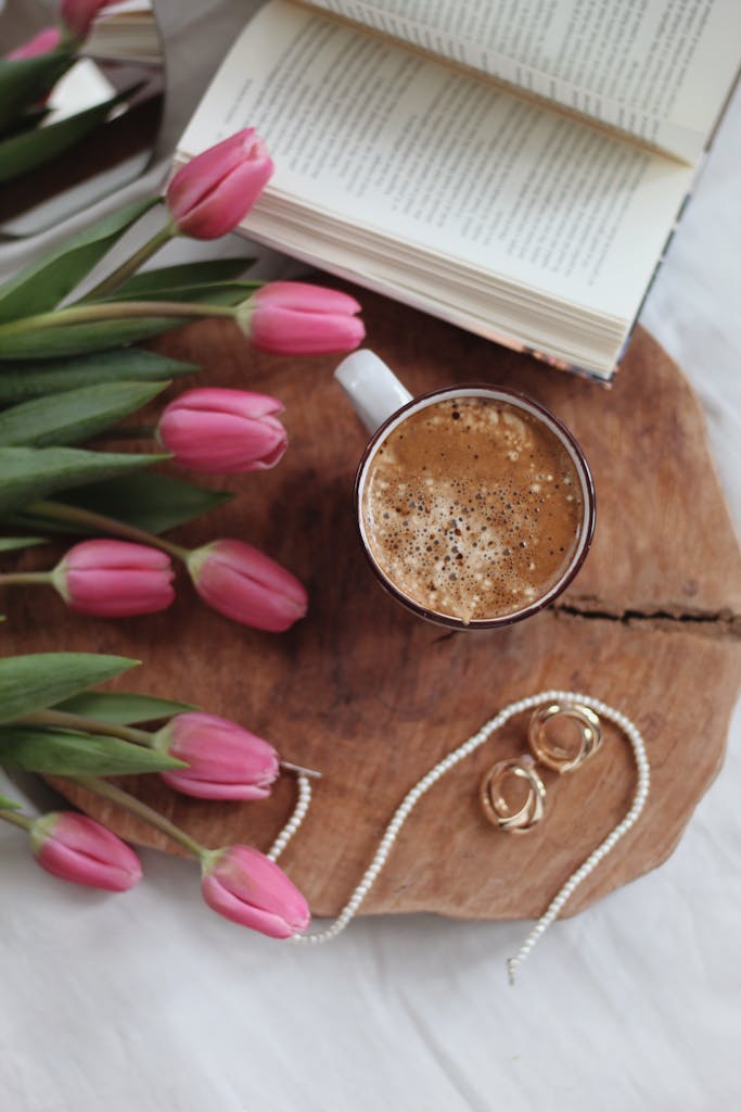 Flat lay of pink tulips, coffee, and book on wooden tray with jewelry.