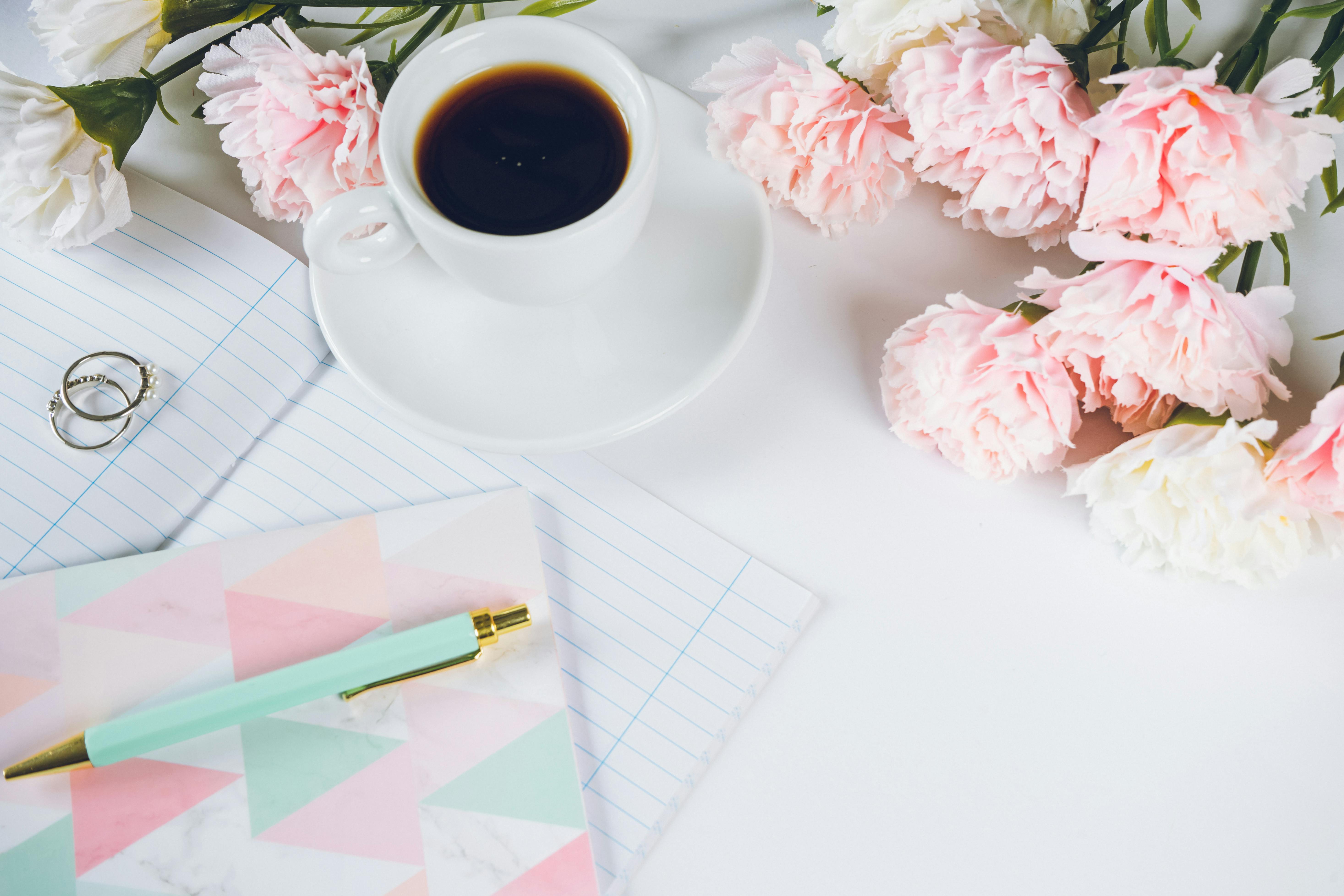 A stylish flat lay with coffee, peonies, notebook, and pen on a white desk.