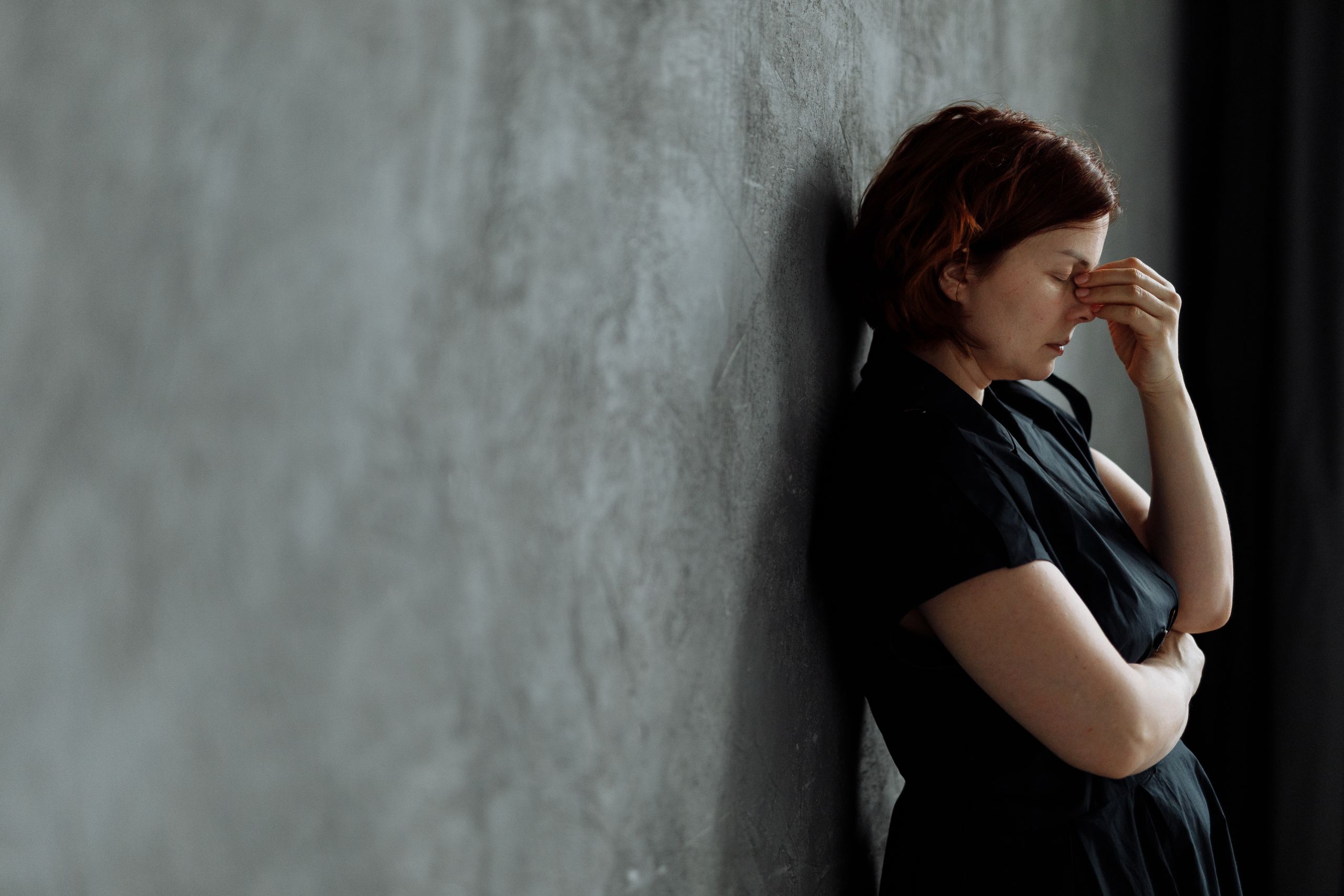 A woman in stress with eyes closed, leaning against a concrete wall indoors.