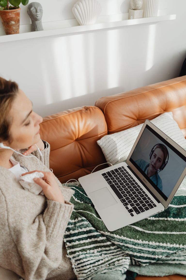 Woman wearing earphones having an online meeting on a laptop, sitting comfortably in a cozy home setting.