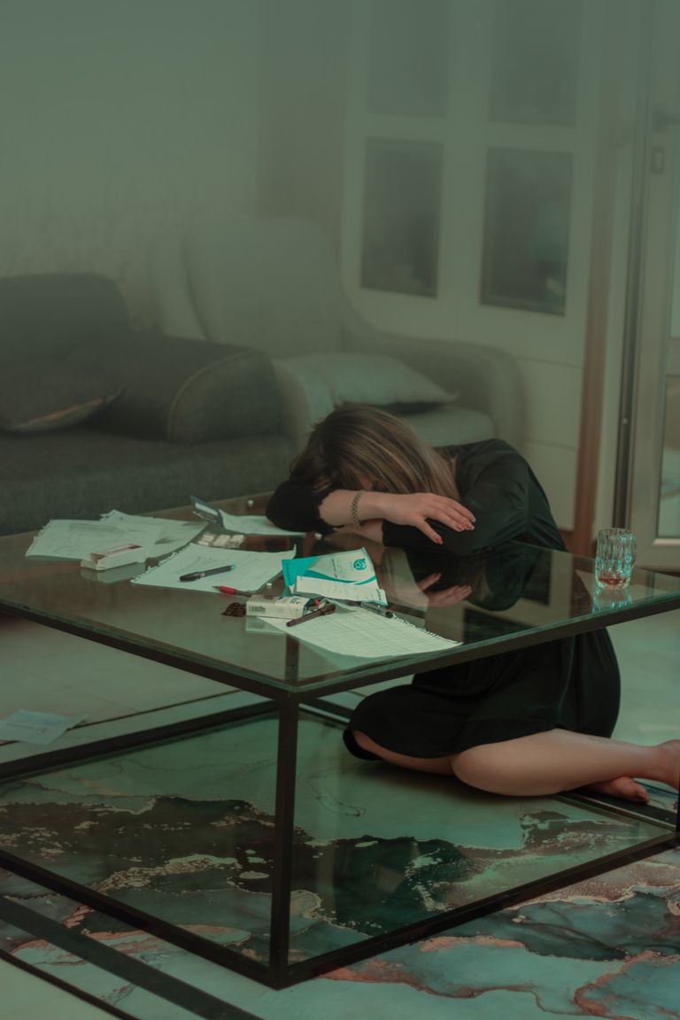 A woman in a bathrobe rests her head on a table filled with scattered bills and documents.