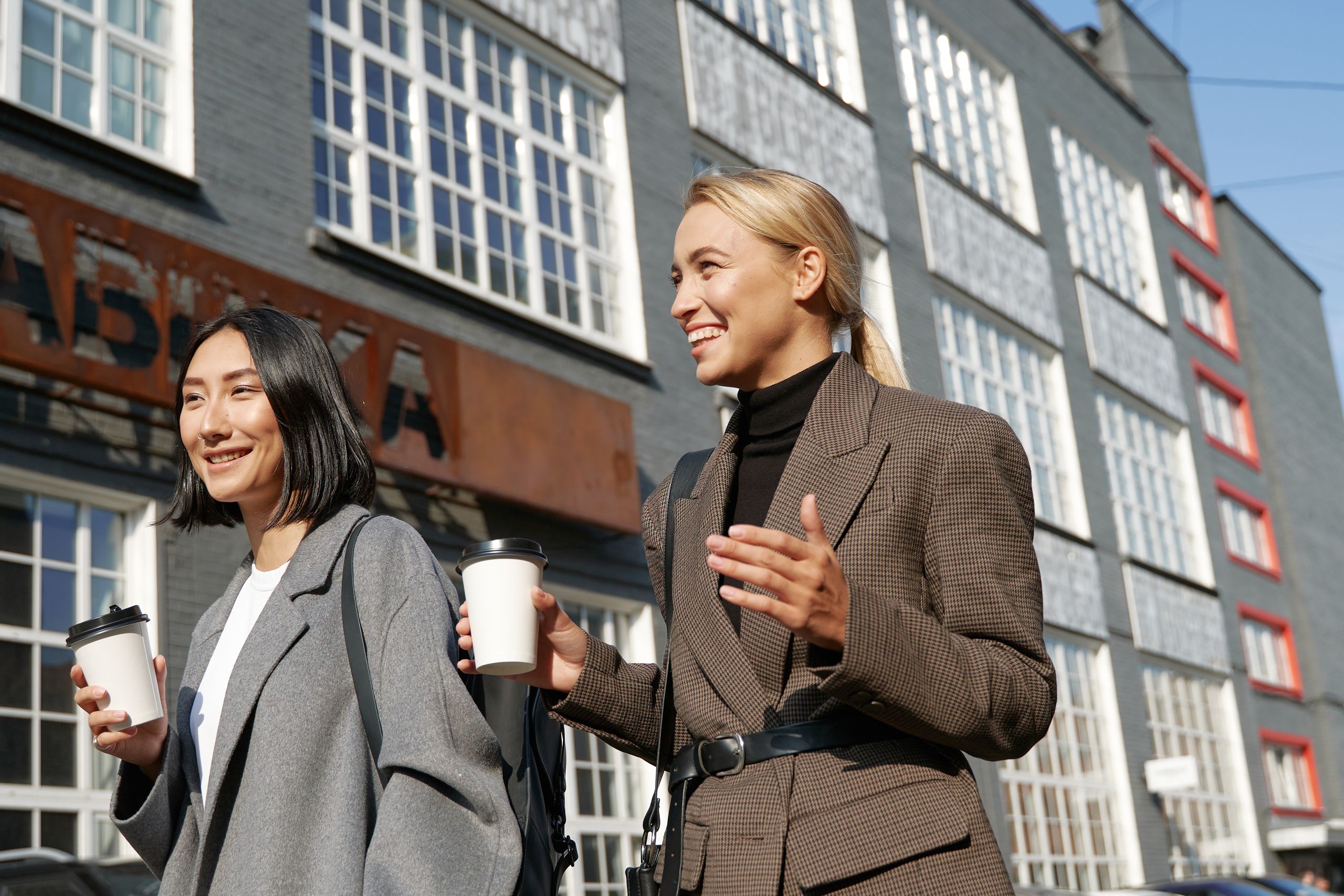 Two women enjoying a sunny day, walking and drinking coffee on a city street.